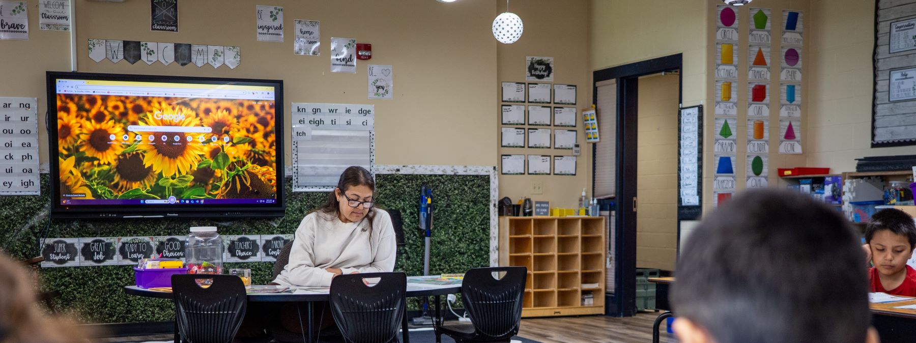 A teacher sits at the front of a classroom, engaging students with a lesson while they listen attentively.
