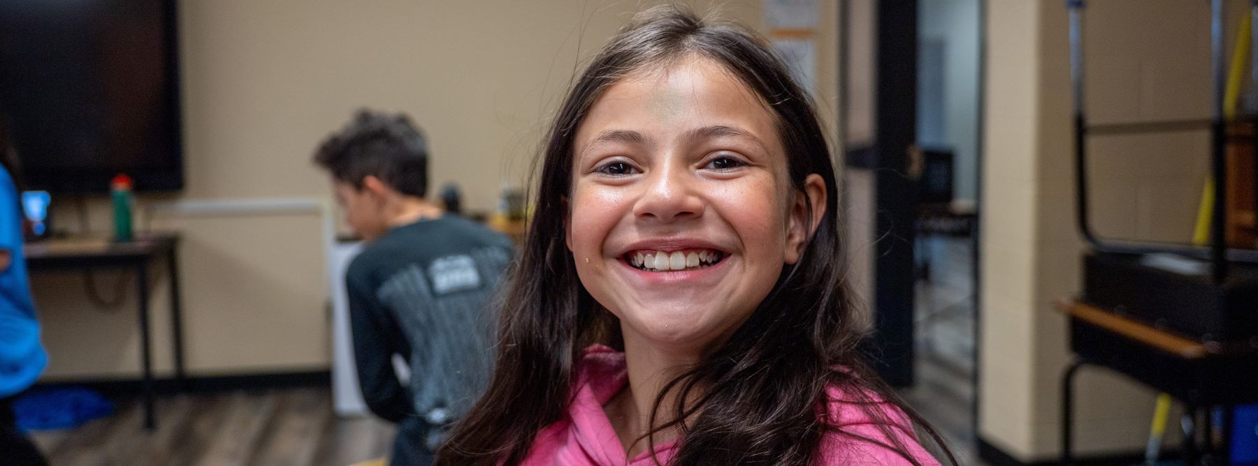 A young girl with a big smile poses for the camera in her classroom, surrounded by colorful posters and desks.