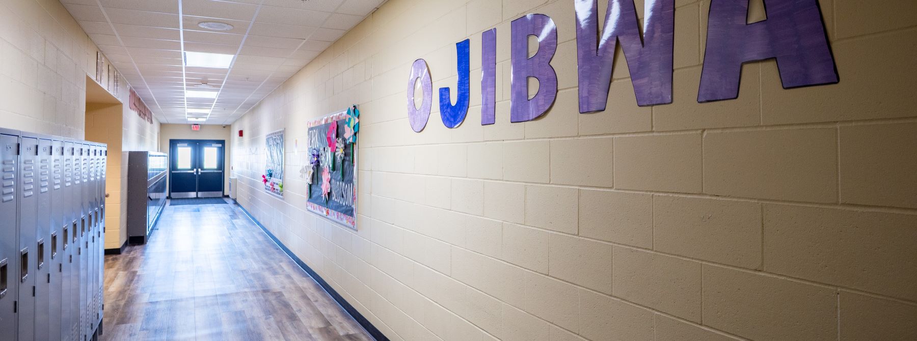 A school hallway lined with lockers, featuring a sign that reads "Ojibwa" prominently displayed on the wall.