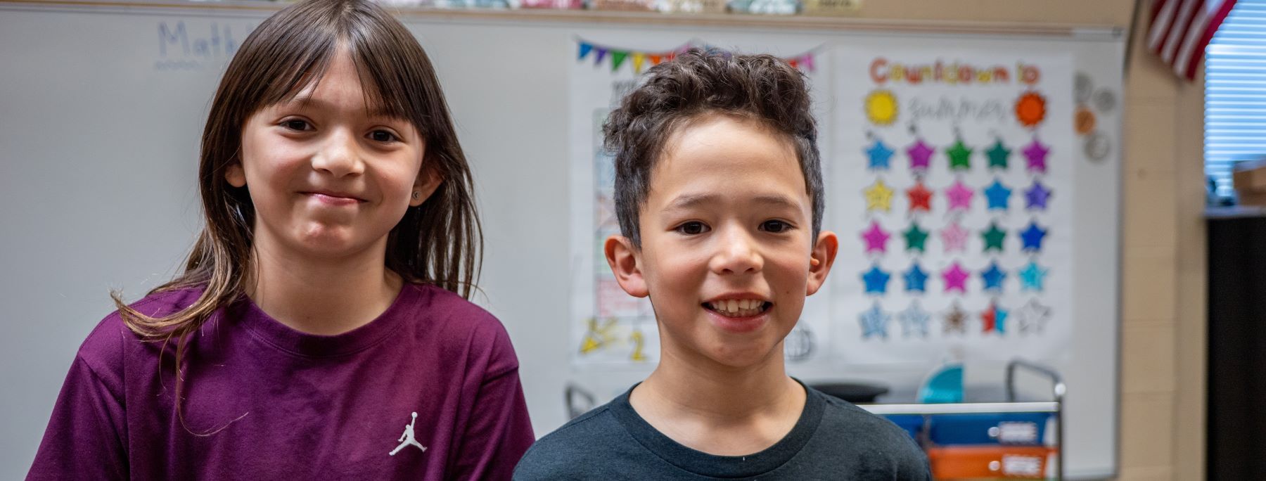 Two smiling students stand in front of a whiteboard, showcasing a joyful learning environment.